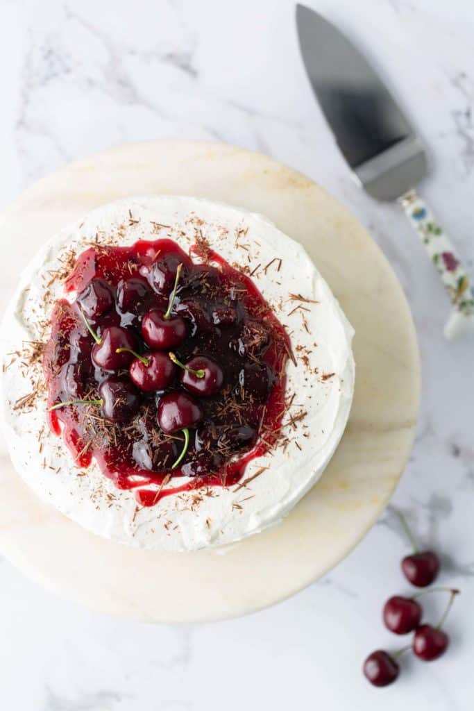 Black Forest Cake A top view of a cake covered with white frosting and topped with cherries and cherry sauce, placed on a marble surface with a serving knife and a few cherries beside the cake.