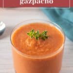A clear glass filled with creamy tomato gazpacho, made from a vibrant gazpacho recipe and garnished with fresh herbs, sits on a light wooden surface.