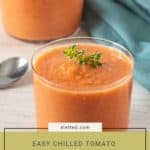 A glass of tomato gazpacho, made from a refreshing gazpacho recipe and garnished with herbs, sits on a table next to a spoon and blue napkin, with another glass in the background.
