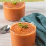 A glass of tomato gazpacho, made from a refreshing gazpacho recipe and garnished with herbs, sits on a wooden surface next to a spoon and green napkin, with another glass in the background.