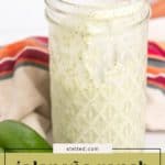A glass jar filled with creamy jalapeño ranch dressing sits in front of a striped cloth, with a fresh jalapeño pepper nearby.