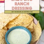 A bowl of creamy jalapeño ranch dressing sits on a plate beside tortilla chips, with fresh jalapeños and a striped cloth in the background.
