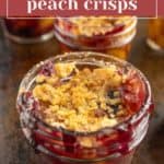 A close-up of a glass jar filled with mini peach crisp, topped with golden crumb topping and blueberries, with more jars of the dessert in the background.