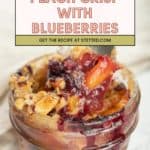 A glass jar filled with homemade peach crisp and juicy blueberries, topped with crumbly oats, with a spoon scooping out a serving. Text overlay reads "Peach Crisp with Blueberries.