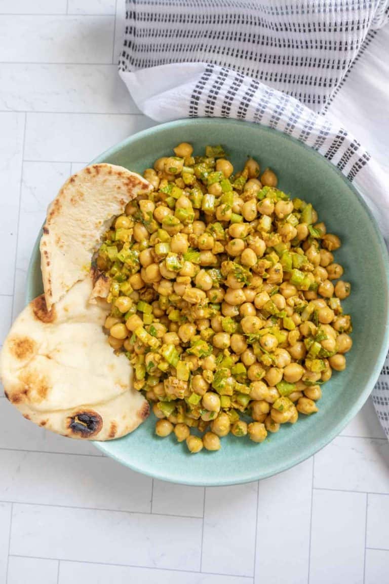 A green bowl filled with chickpea salad containing diced vegetables, accompanied by two pieces of naan bread on the side. A white and black patterned cloth napkin is partially visible.