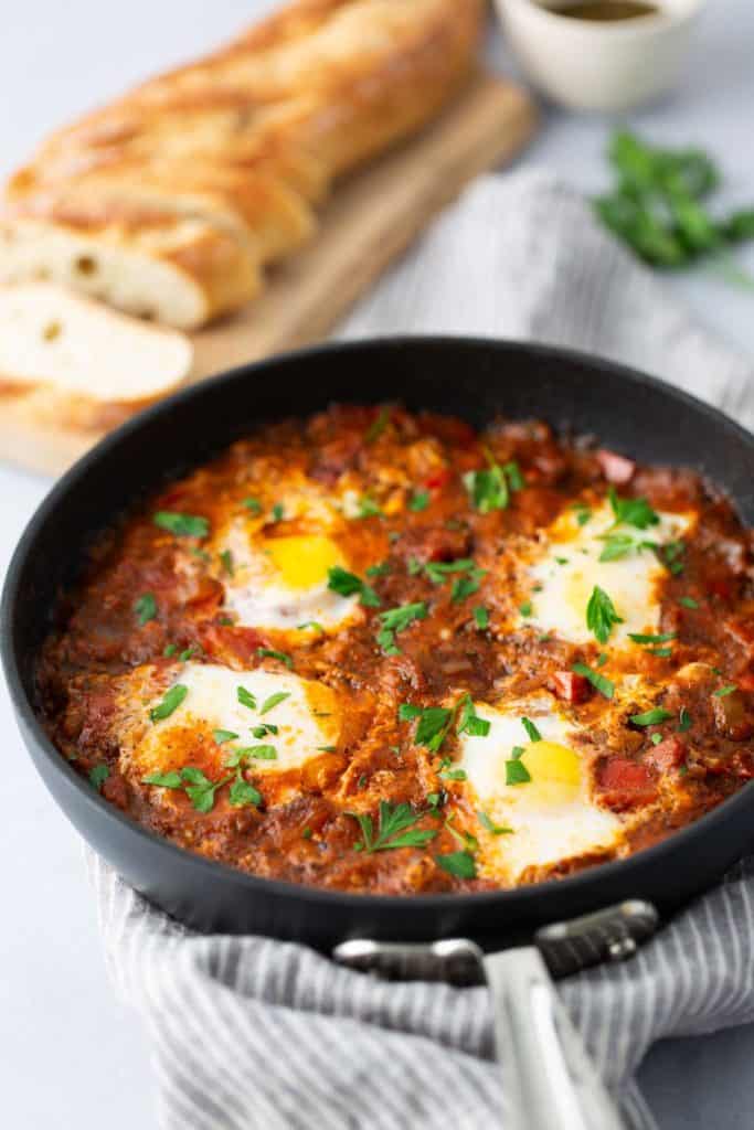 A skillet with shakshuka, a dish of poached eggs in a tomato and pepper sauce, garnished with parsley. Slices of bread and a bowl of sauce are in the background.