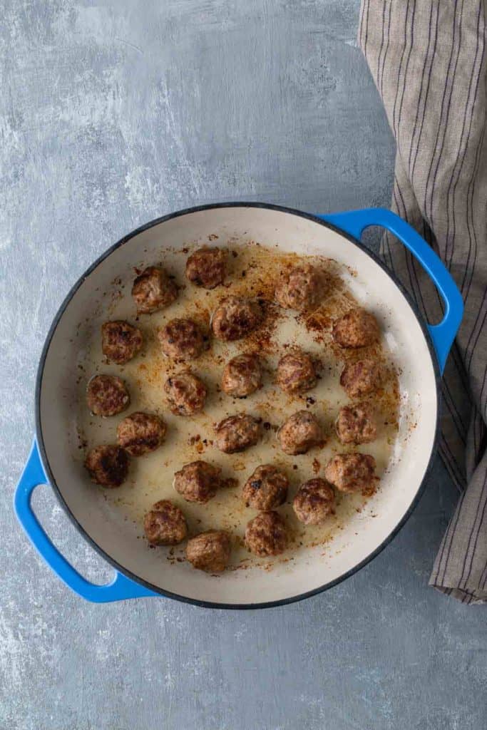 A blue-handled baking dish containing cooked meatballs sits on a gray surface next to a gray-striped cloth.