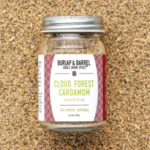 A glass jar of Burlap & Barrel Cloud Forest ground cardamom sits on a bed of cardamom seeds. The label indicates this ground spice is sourced from Alta Verapaz, Guatemala.