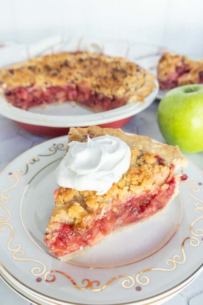 Apple Cranberry Pie A slice of crumble-topped fruit pie with whipped cream on a decorative plate, next to a whole pie with a slice missing, and a green apple on the side.