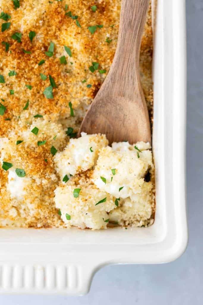 Cauliflower Gratin A white baking dish with a wooden spoon scooping into a cauliflower and breadcrumb casserole, garnished with chopped herbs.