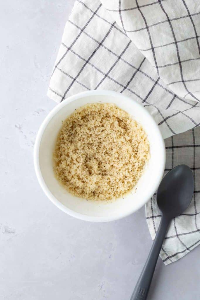 A bowl of breadcrumbs on a light surface, next to a black spoon and a checkered cloth.