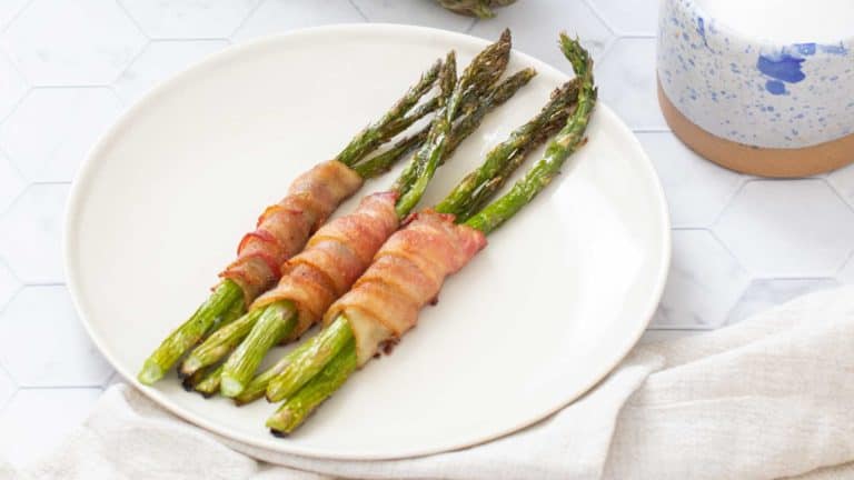 Three bacon-wrapped asparagus bundles on a white plate, placed on a hexagonal tile surface.