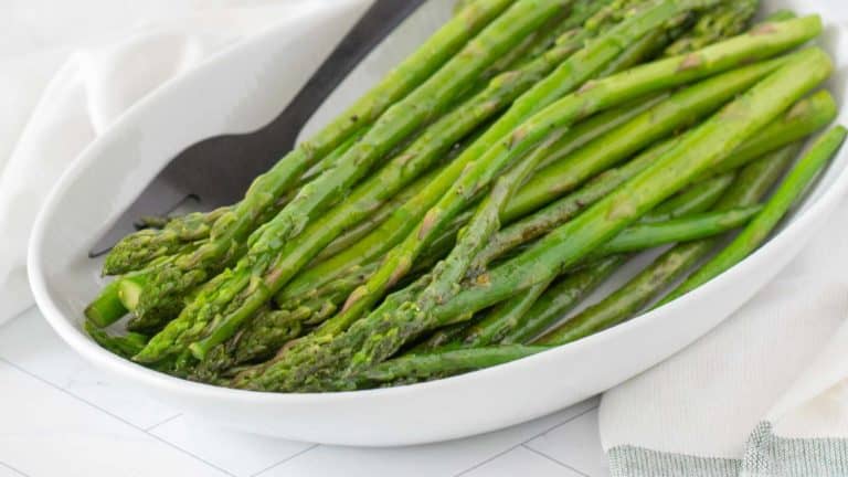 A white oval dish with cooked green asparagus spears and a black serving fork.