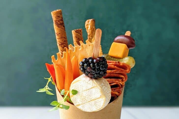 A paper cup filled with cheese, breadsticks, sliced vegetables, a blackberry, and skewered meats and cheeses, arranged in an upright, decorative manner against a green background.