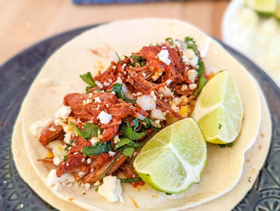A soft taco filled with shredded meat, crumbled cheese, chopped onions, cilantro, and two lime wedges on a plate.