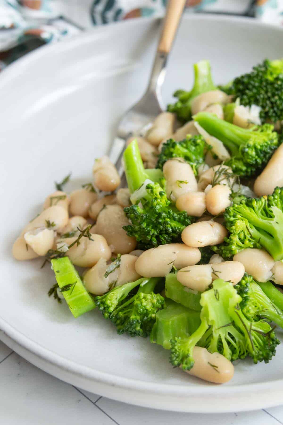 A white plate with cooked white beans and broccoli florets, garnished with herbs, with a fork resting on the side.