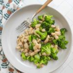 A plate of cooked broccoli and white beans with herbs, served with a fork on a light-colored surface next to a patterned cloth.