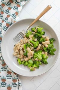A plate of cooked broccoli and white beans with herbs, served with a fork on a light-colored surface next to a patterned cloth.