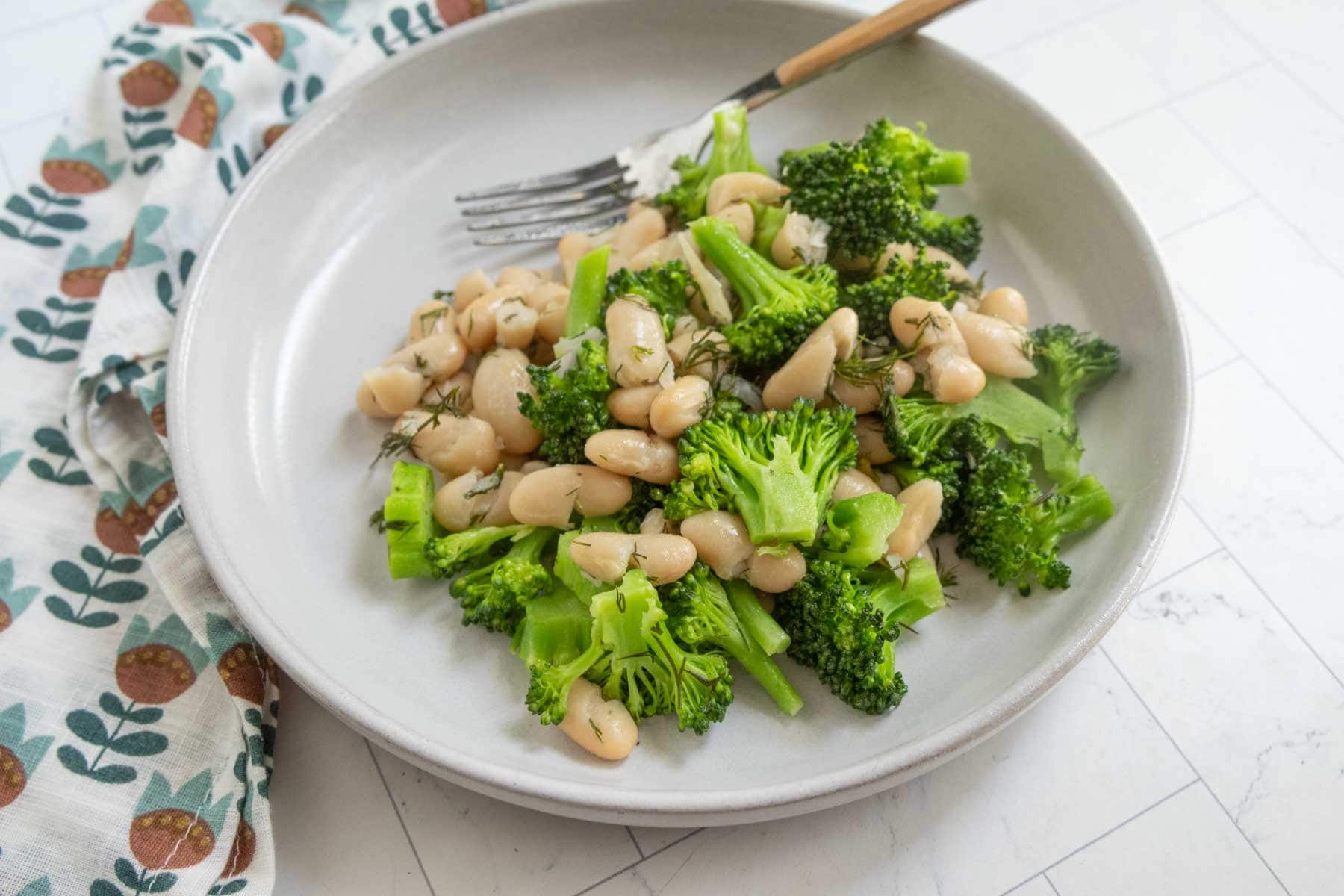 A plate of steamed broccoli and white beans, garnished with herbs, with a fork resting on the side.