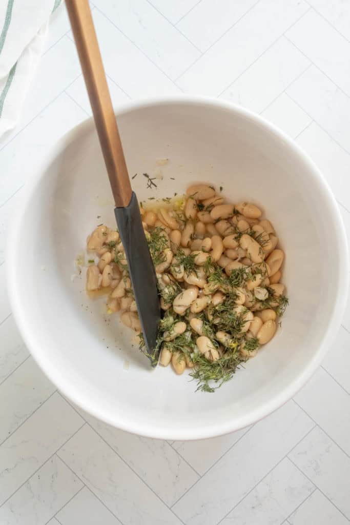 A white bowl containing white beans, chopped herbs, and a black spatula, placed on a light-colored tile surface.