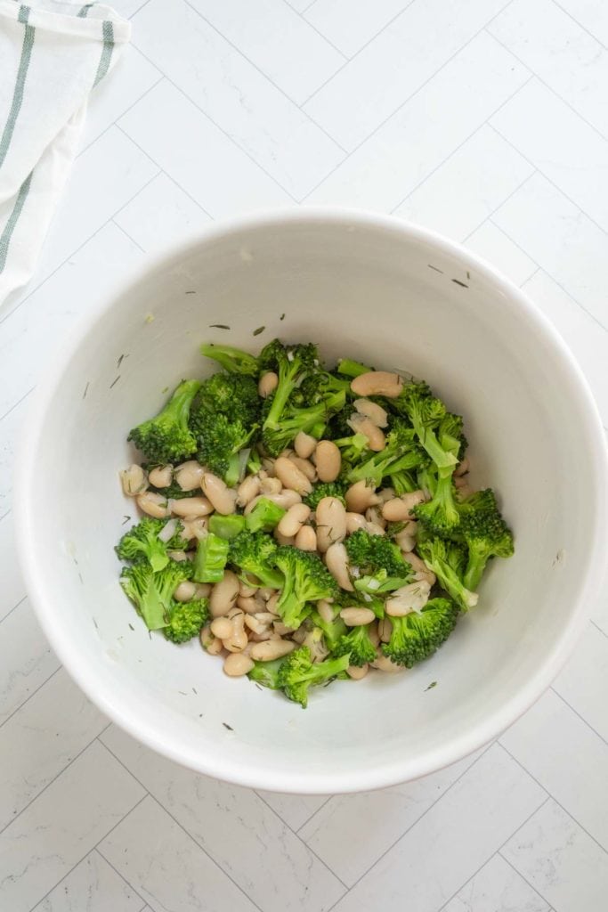 A white bowl containing cooked broccoli florets and white beans, placed on a white tiled surface.