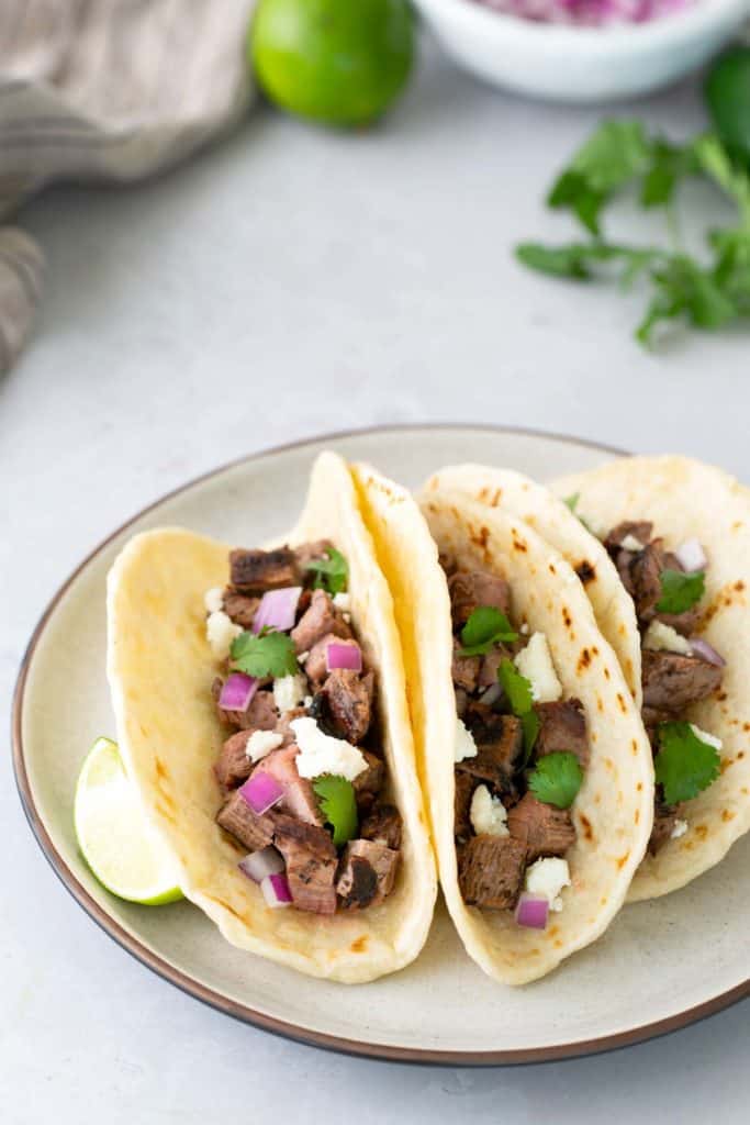 Three soft flour tortillas filled with grilled steak, chopped red onion, fresh cilantro, and crumbled cheese are served on a plate with lime wedges.