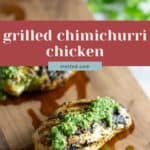 Two pieces of grilled chicken breast topped with green chimichurri sauce on a wooden cutting board, with chopped herbs in the background.