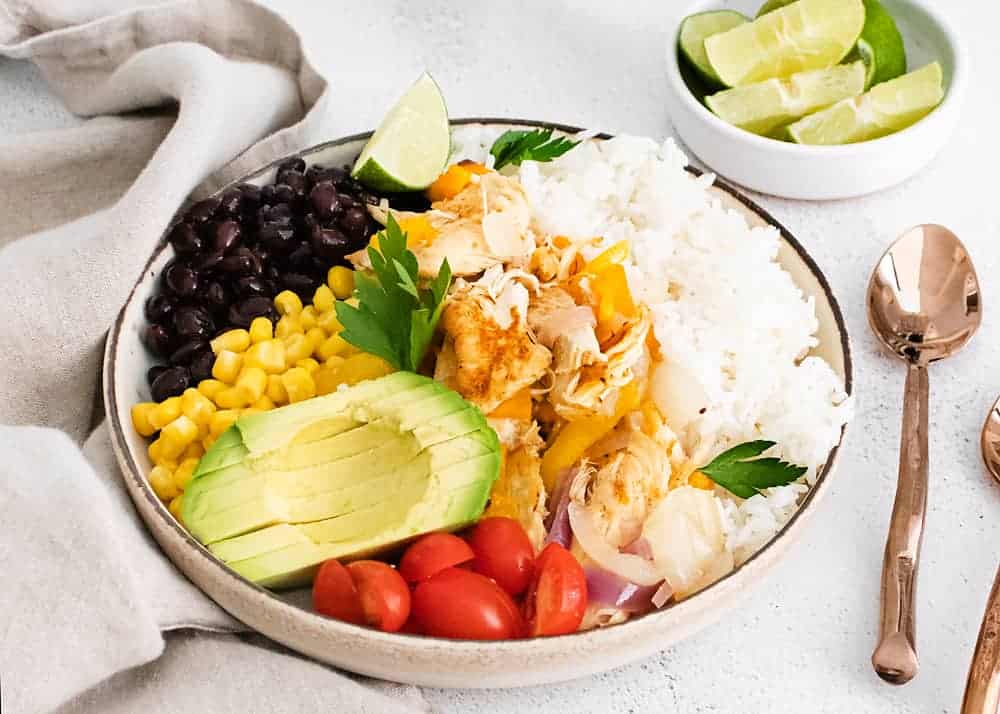 A bowl with white rice, shredded chicken, black beans, corn, avocado slices, cherry tomatoes, and herbs. A small bowl of lime wedges and a spoon are beside it.