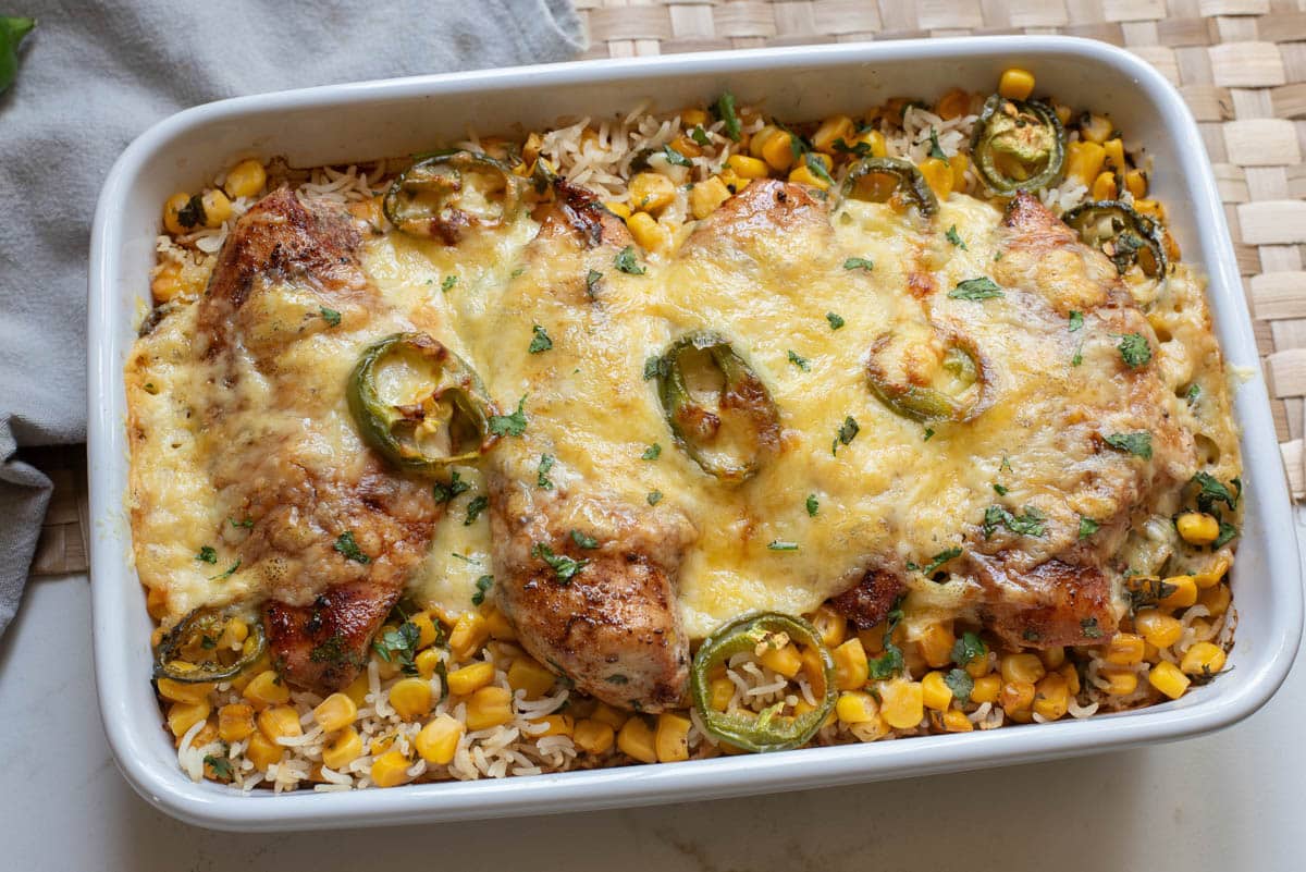 A baked casserole dish with chicken breasts, rice, corn, melted cheese, and jalapeño slices, garnished with chopped herbs.