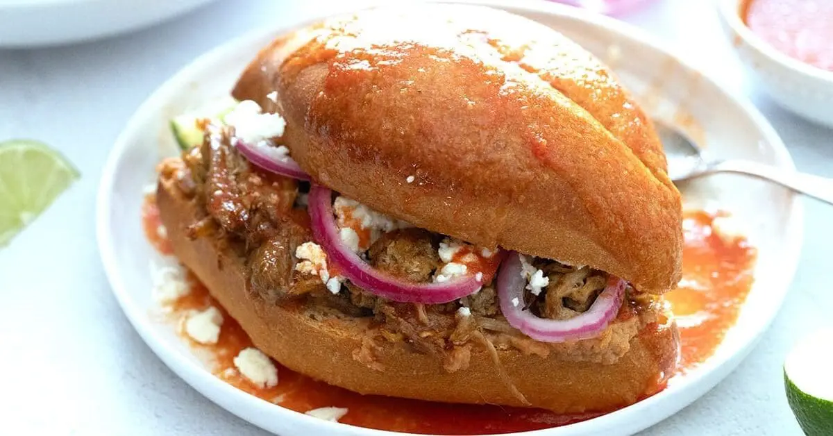 A Mexican torta ahogada sandwich filled with shredded meat, pickled red onions, and cheese, served on a white plate with red sauce.