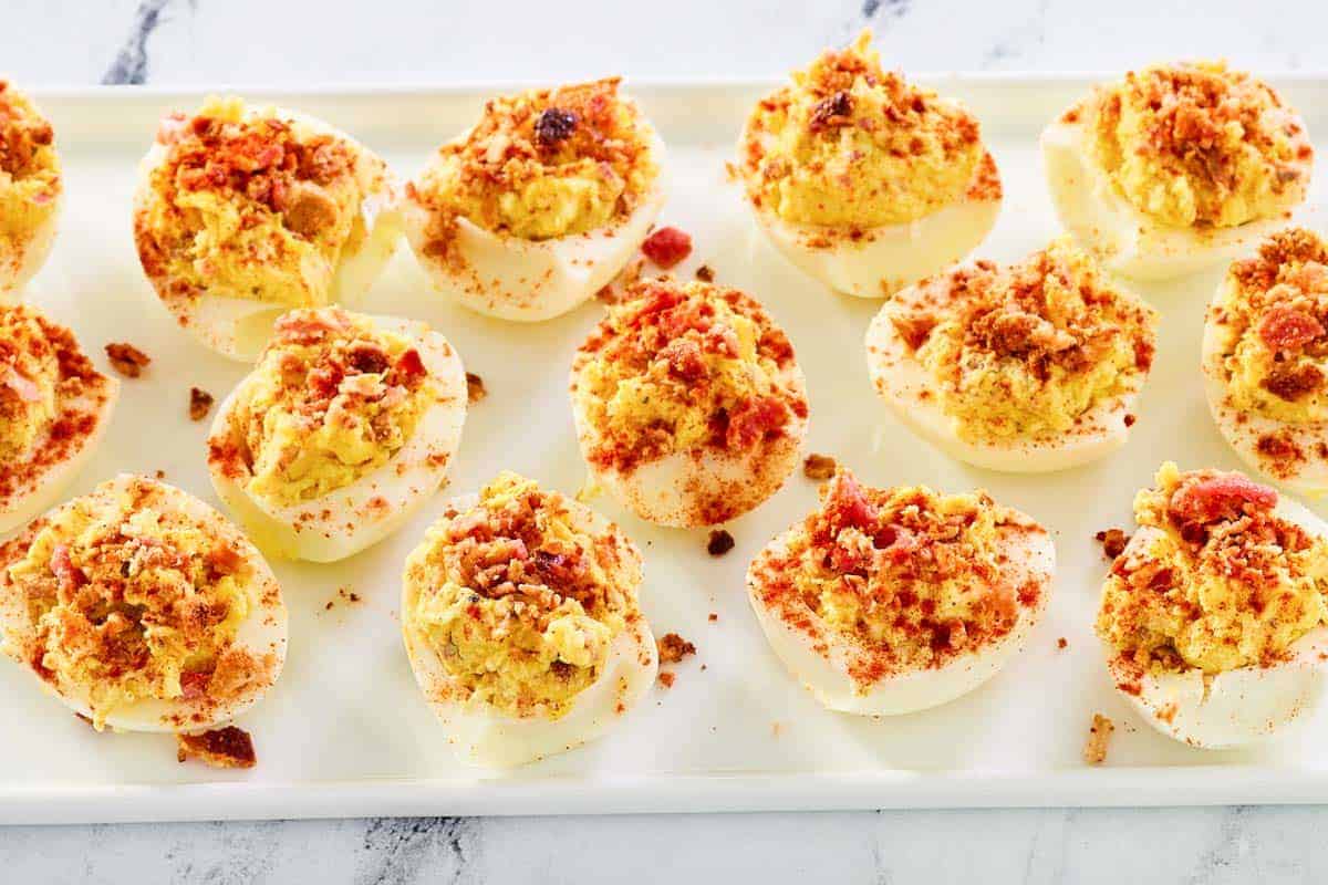 A tray of deviled eggs topped with crumbled bacon and seasoning, arranged in rows on a white rectangular plate.