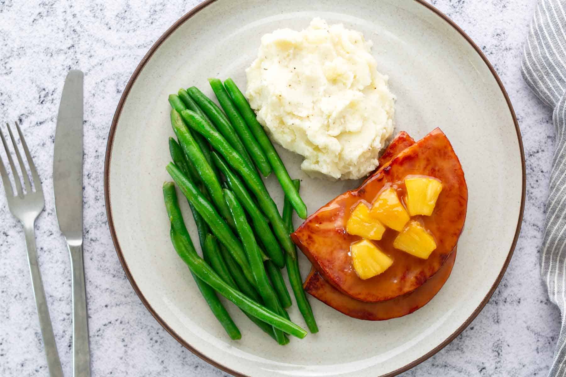 A plate with green beans, mashed potatoes, and glazed ham topped with pineapple chunks. Knife and fork on the side.