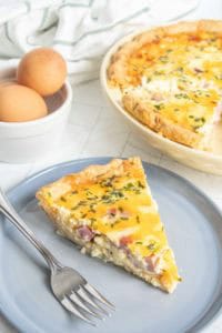 A slice of quiche with ham and herbs on a gray plate, accompanied by a fork. A whole quiche and a bowl with two eggs are in the background.