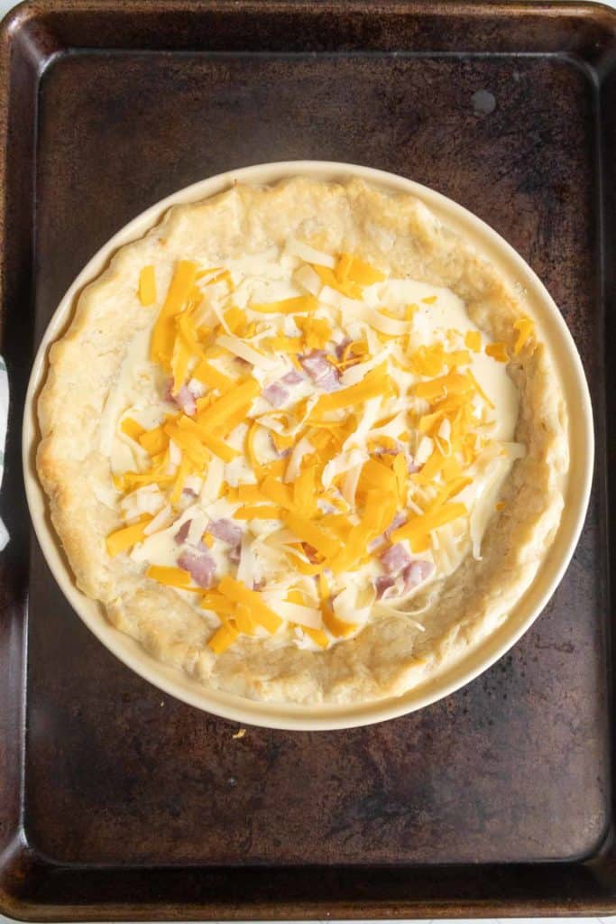 Unbaked quiche in a round dish on a baking tray, filled with shredded cheese and diced ham on a creamy mixture, with a flaky crust.