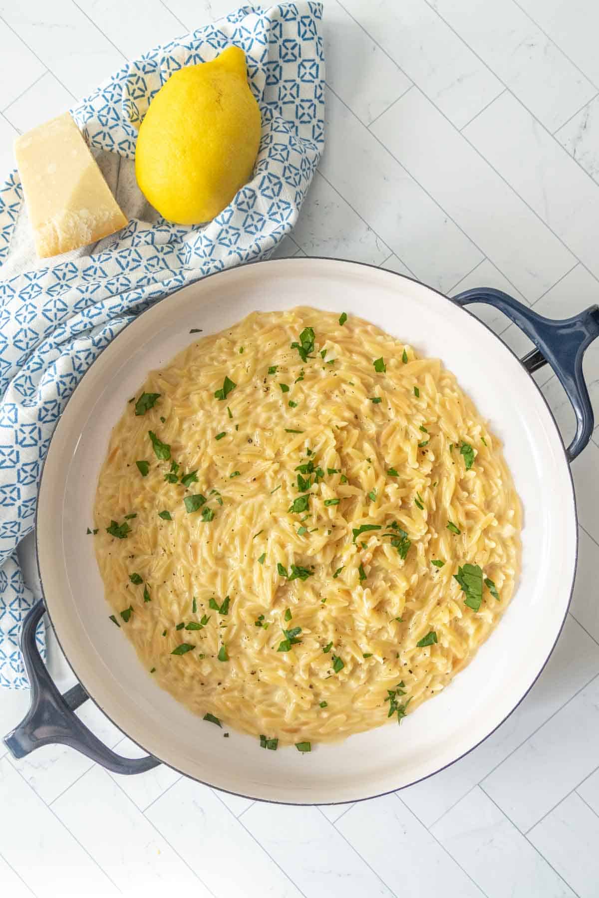 A white pan filled with creamy orzo pasta topped with chopped herbs, placed on a white table near a lemon and a chunk of parmesan cheese on a patterned blue cloth.