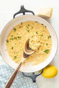 A pan of creamy orzo pasta garnished with chopped herbs, with a wooden spoon, a piece of cheese, and a lemon placed nearby.