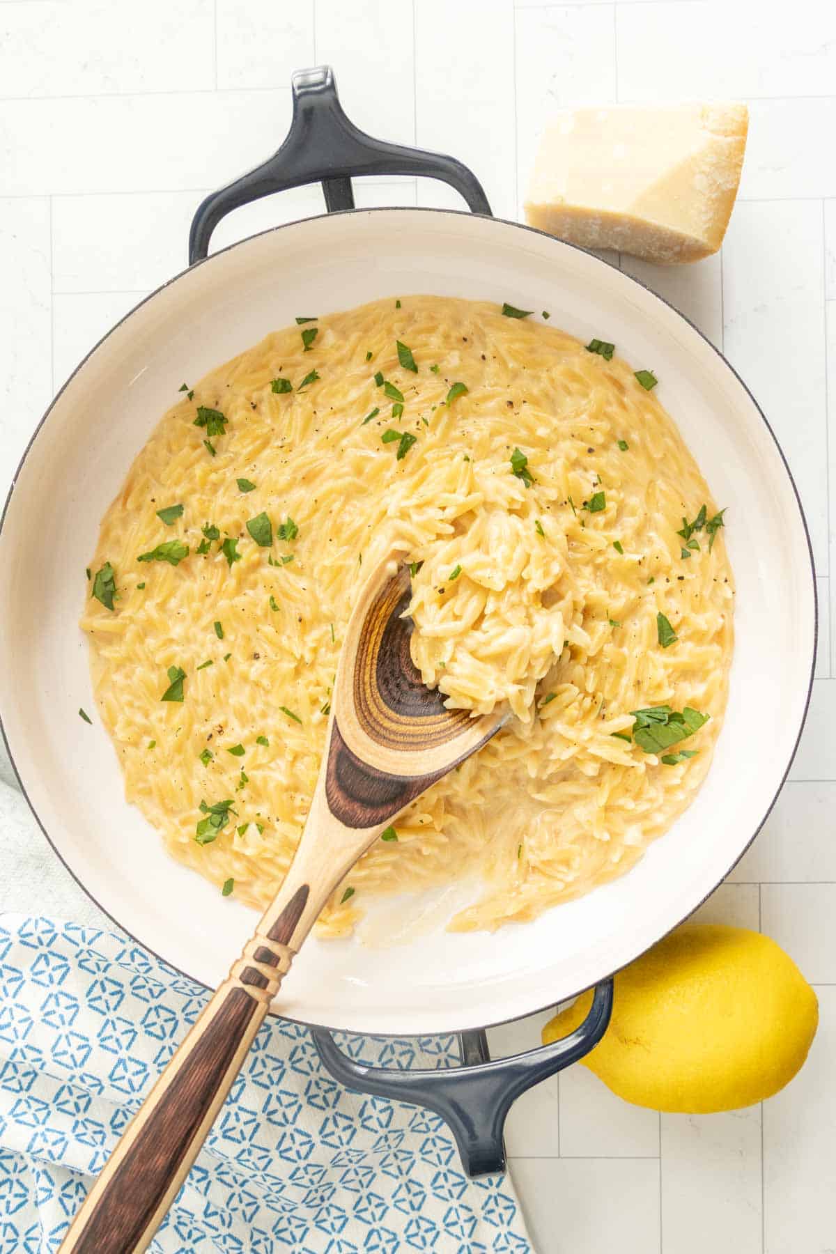 A pan of creamy orzo pasta garnished with chopped herbs, with a wooden spoon, a piece of cheese, and a lemon placed nearby.