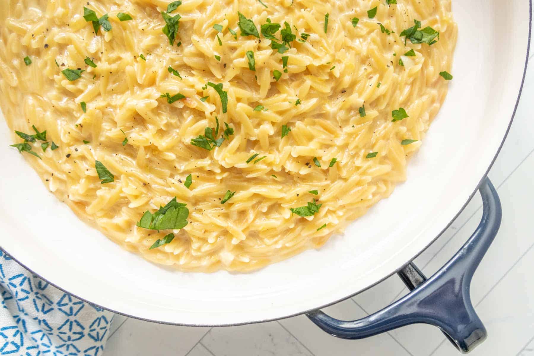 A white pan filled with creamy orzo pasta topped with chopped parsley, placed on a white surface next to a blue-patterned cloth.
