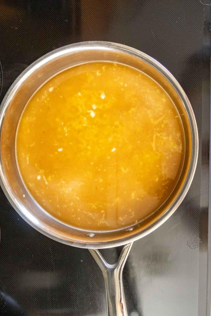 A stainless steel saucepan on a stovetop contains a light brown broth with small pieces of garlic or onion visible.