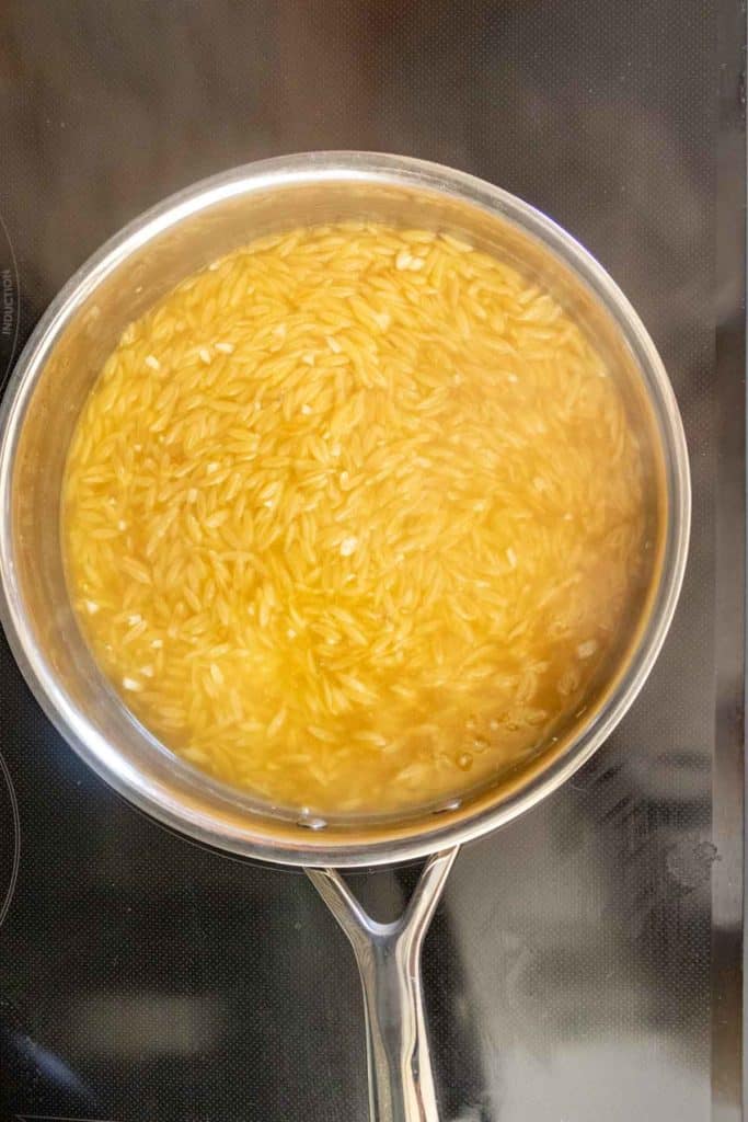 A stainless steel pot filled with orzo pasta cooking in simmering water on a black stovetop.