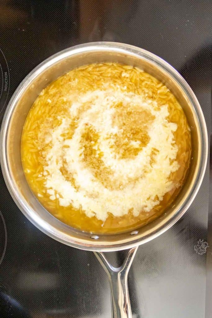 A saucepan on a stove containing orzo pasta cooking in broth, with grated cheese sprinkled on top.