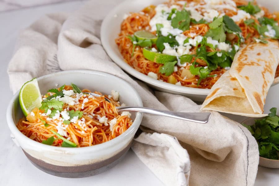 A bowl and plate of Mexican fideo topped with cheese, cilantro, and lime, served with a flour tortilla on a cloth napkin.