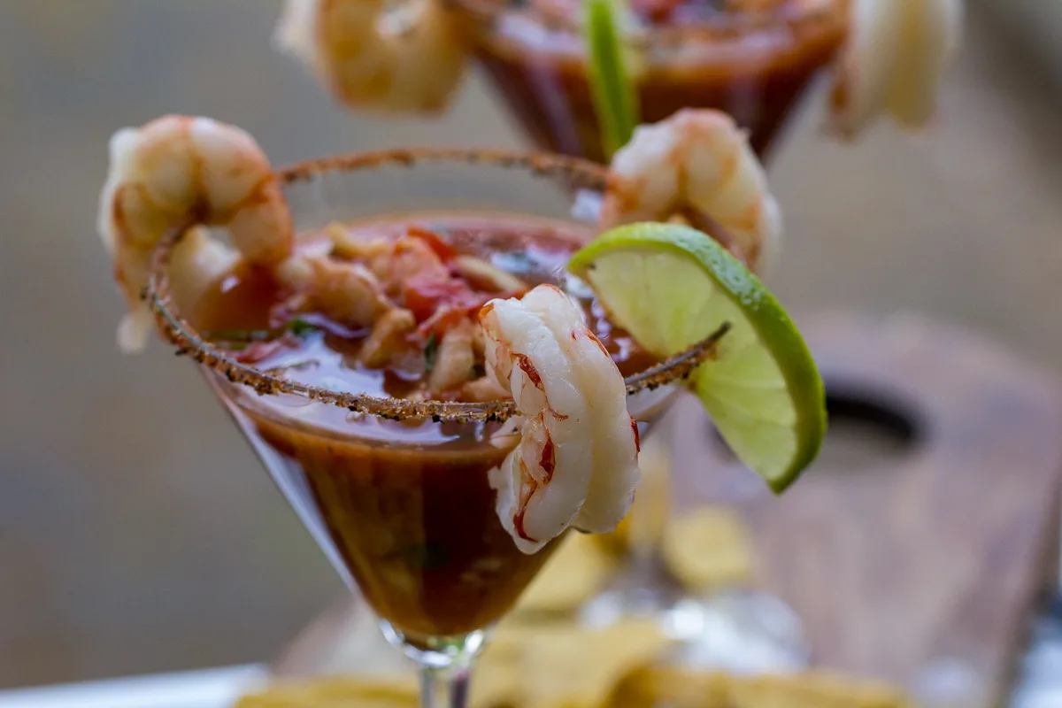 A shrimp cocktail in a martini glass garnished with shrimp, a lime slice, and a spiced rim, with tortilla chips in the background.