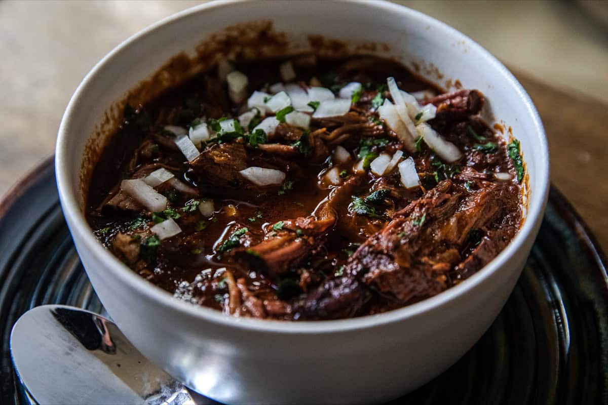 A white bowl of beef stew topped with chopped onions and herbs, placed on a dark plate with a spoon beside it.