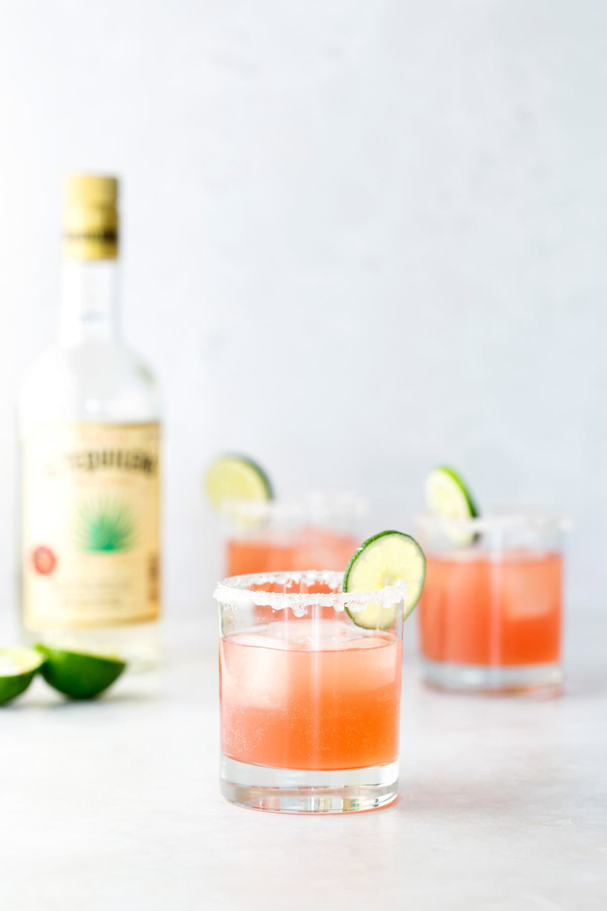 Three pink cocktails with salted rims and lime slices are in the foreground, with a bottle of tequila and lime wedges in the background on a light surface.