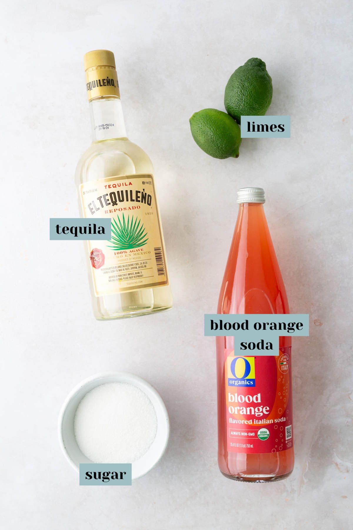A bottle of tequila, two limes, a bottle of blood orange soda, and a bowl of sugar are arranged on a light surface, each labeled with its name.