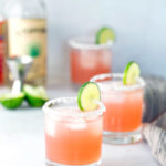 Three glasses of pink cocktail with salted rims and lime slices, ice, a bottle of liquor, lime wedges, and a cocktail jigger are visible in the background.