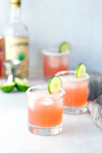 Three glasses of pink cocktail with salted rims and lime slices, ice, a bottle of liquor, lime wedges, and a cocktail jigger are visible in the background.