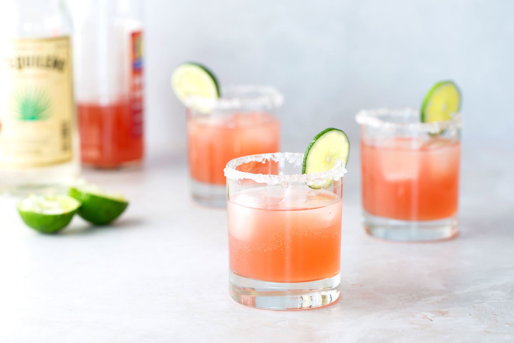 Three glasses of pink cocktail with salted rims and cucumber slices, placed on a light surface with bottles and lime halves in the background.