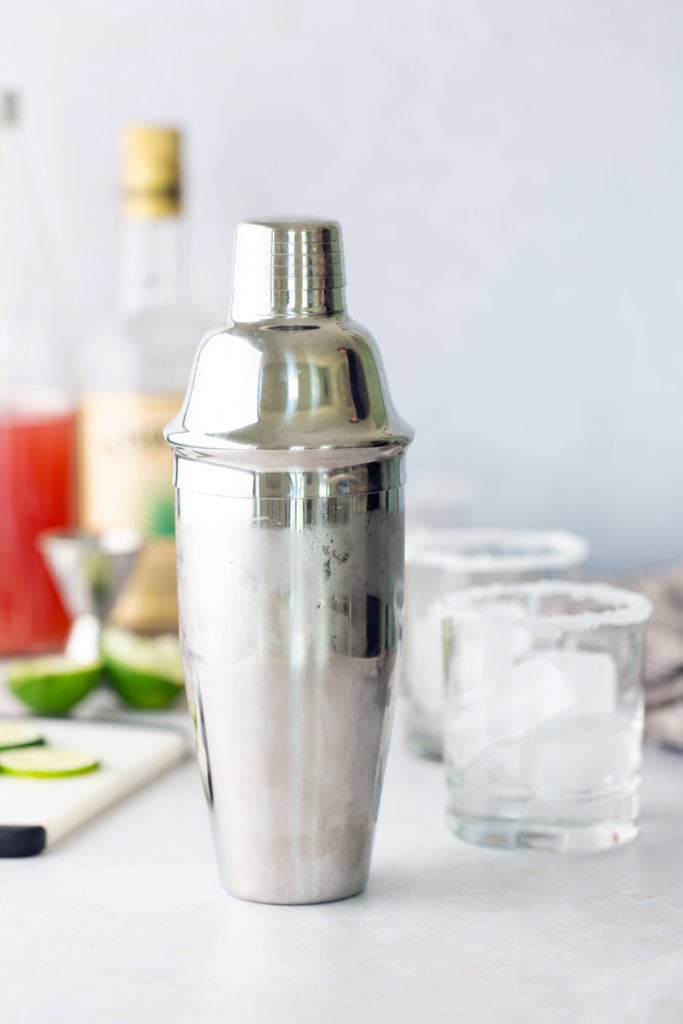 A stainless steel cocktail shaker is placed on a countertop with two glasses, a lime wedge, and bottles blurred in the background.